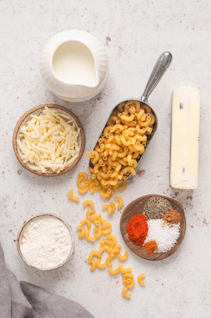 Ingredients on a textured background, including elbow noodles, shredded cheddar, butter, flour, milk and spices.