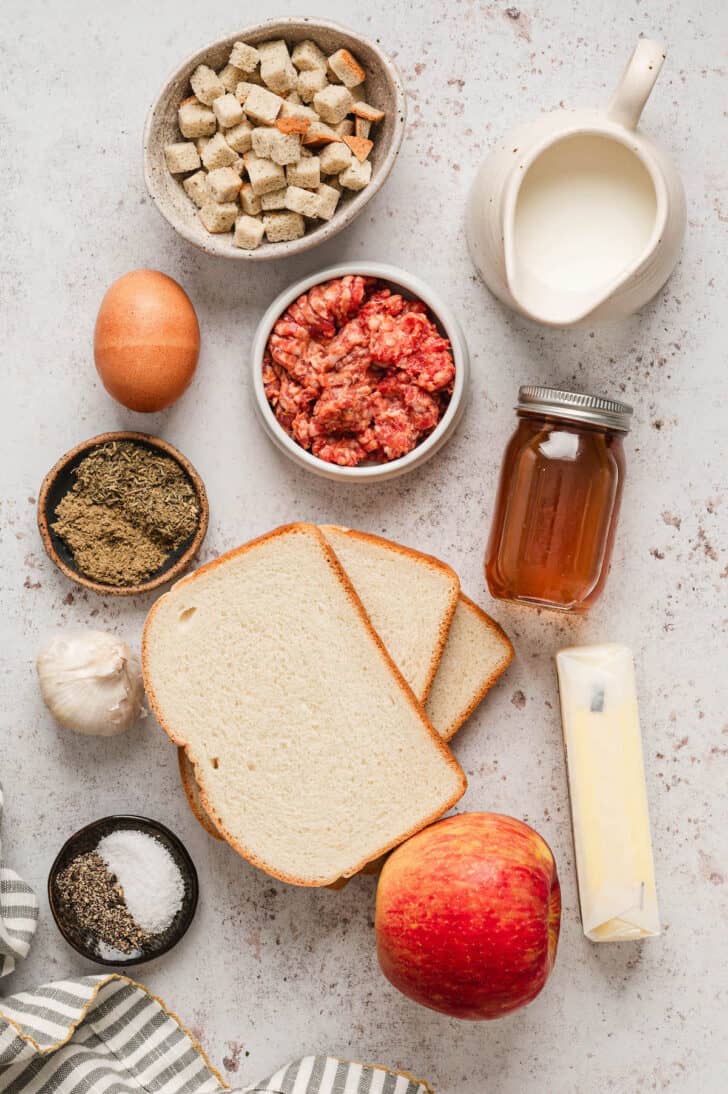 Ingredients on a light surface, including bread, bread cubes, raw ground meat, broth, butter, milk, garlic, apple, egg, herbs and salt and pepper.