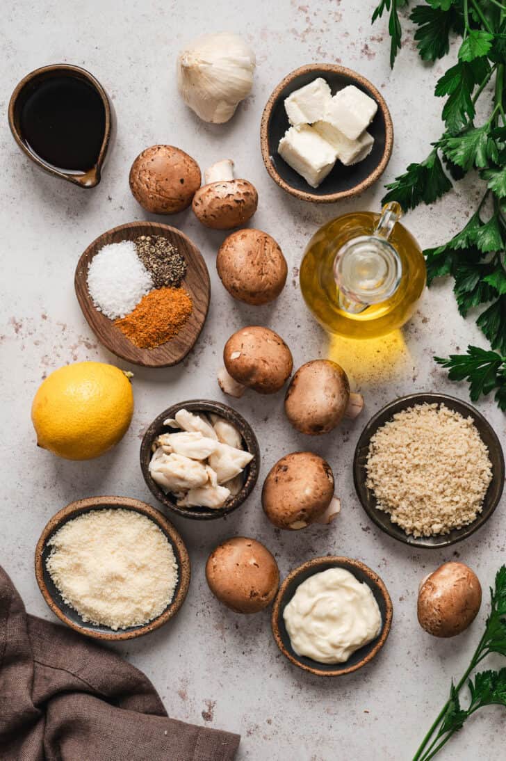 Ingredients on a light surface, including creminis, cream cheese, mayonnaise, lemon, herbs, cheese and breadcrumbs.