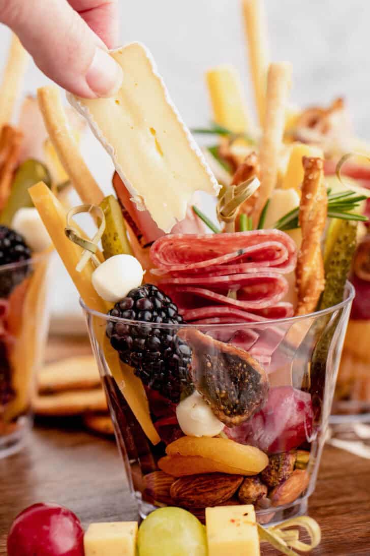 A hand removing a piece of brie from a small glass filled with snacks.