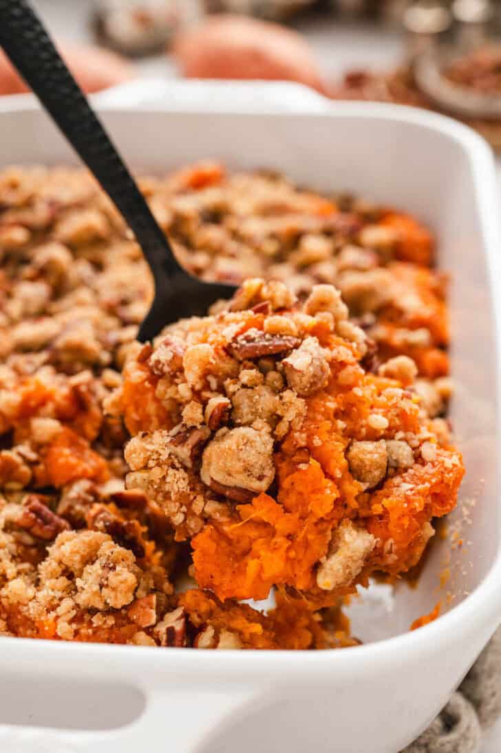 A black spoon lifting a scoop of easy sweet potato casserole out of a white baking dish.