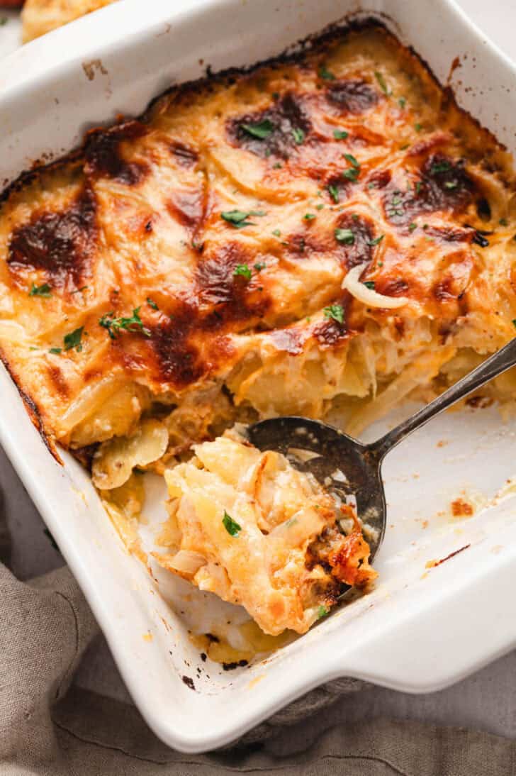 A spoon lifting a scoop of cheesy scalloped potatoes from a white baking dish.