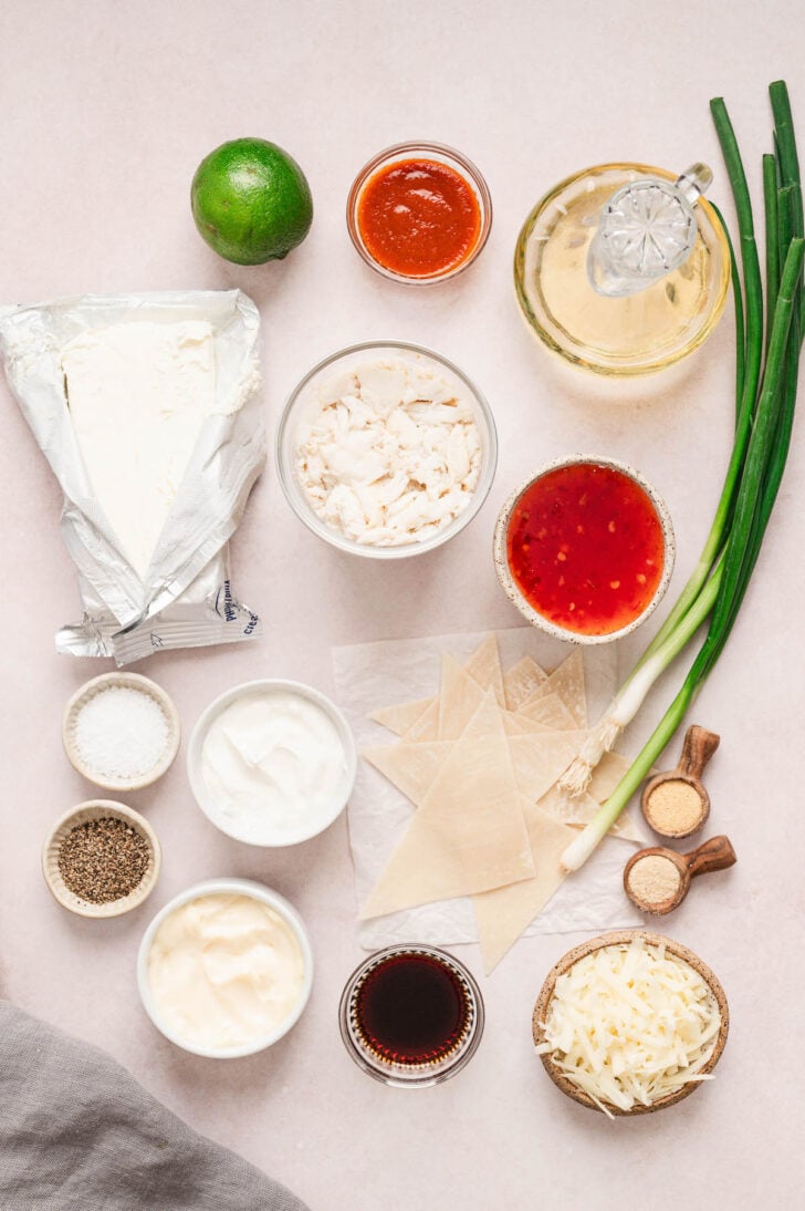 Ingredients on a light surface, including seafood, cheese, mayonnaise, cream cheese, spices, green onions, soy sauce, wonton wrappers and sweet chili sauce.