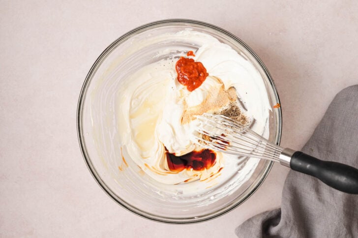 A creamy mixture topped with spices, hot sauce and soy sauce in a glass bowl with a whisk.