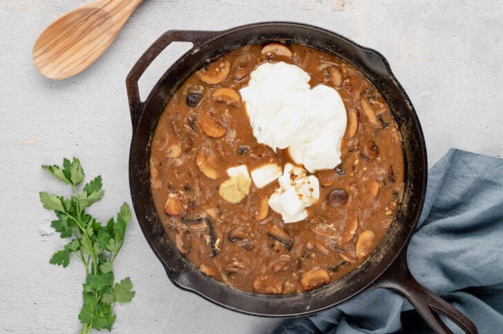 Sour cream being added to a brown saucy mushroom mixture in a skillet.