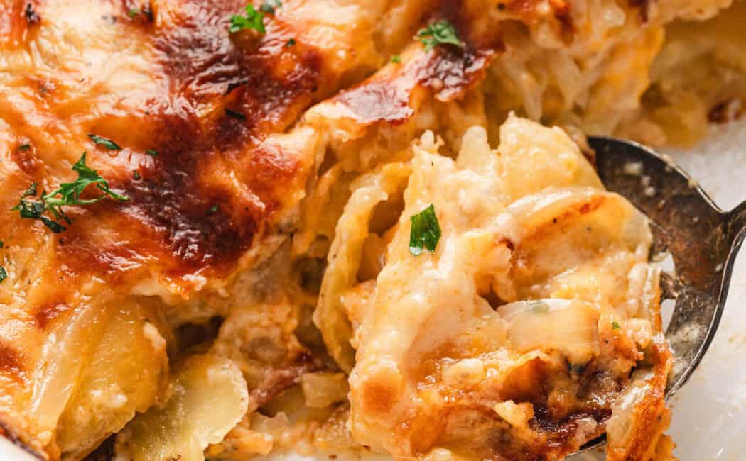 A spoon lifting a scoop of cheesy scalloped potatoes from a white baking dish.