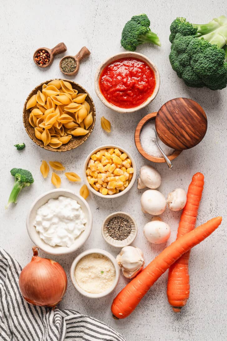 Ingredients on a light surface, including carrots, mushrooms, cottage cheese, onion, marinara, corn, spices, broccoli and shells.