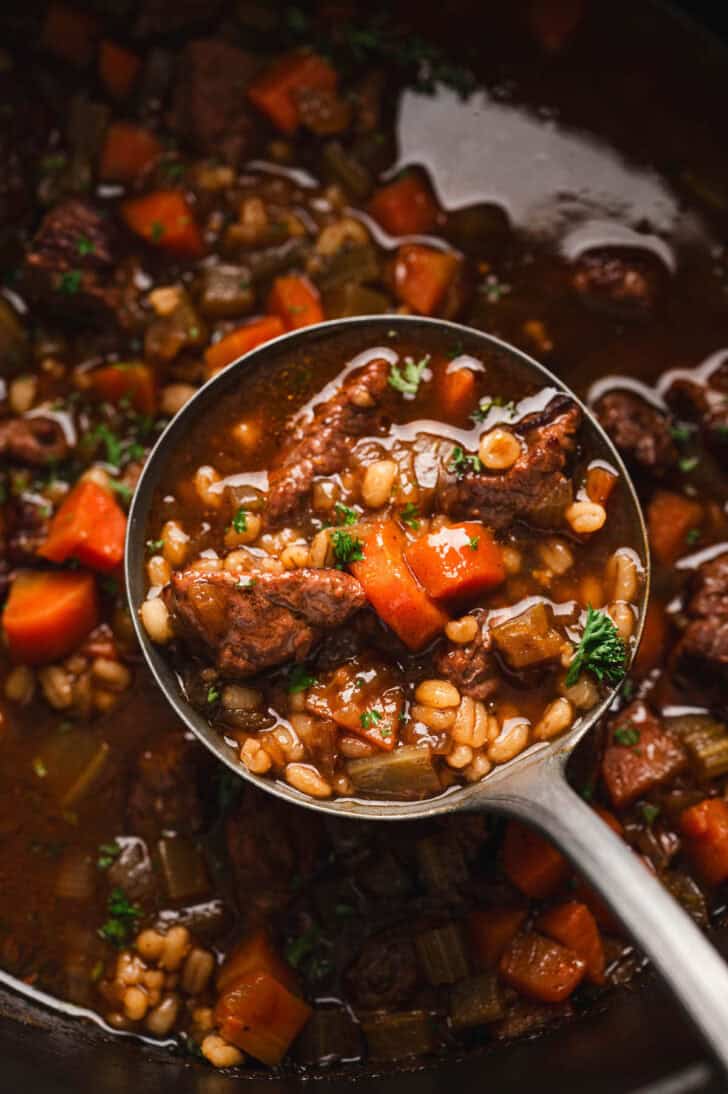 A ladle scooping a serving of the best beef barley soup recipe from a pot.