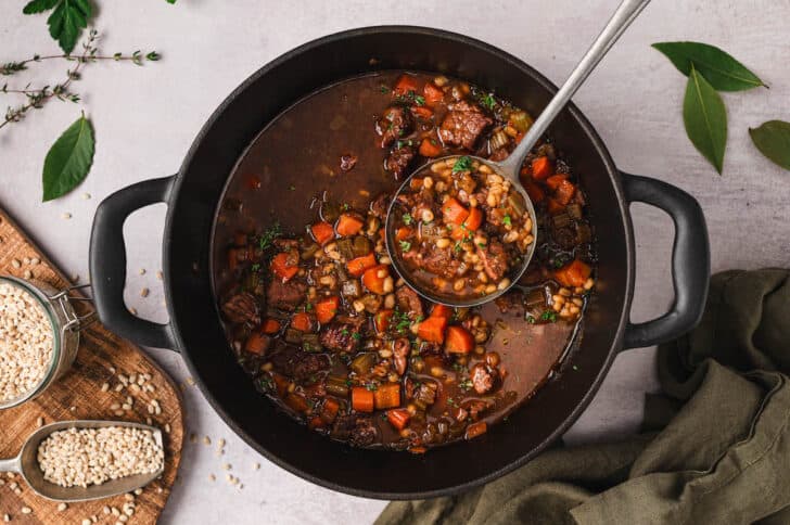 A ladle scooping beef and barley soup from a Dutch oven.