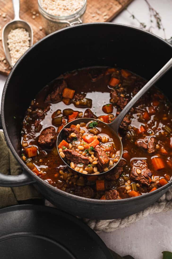 A ladle scooping a serving of a beef and barley soup recipe from a Dutch oven.