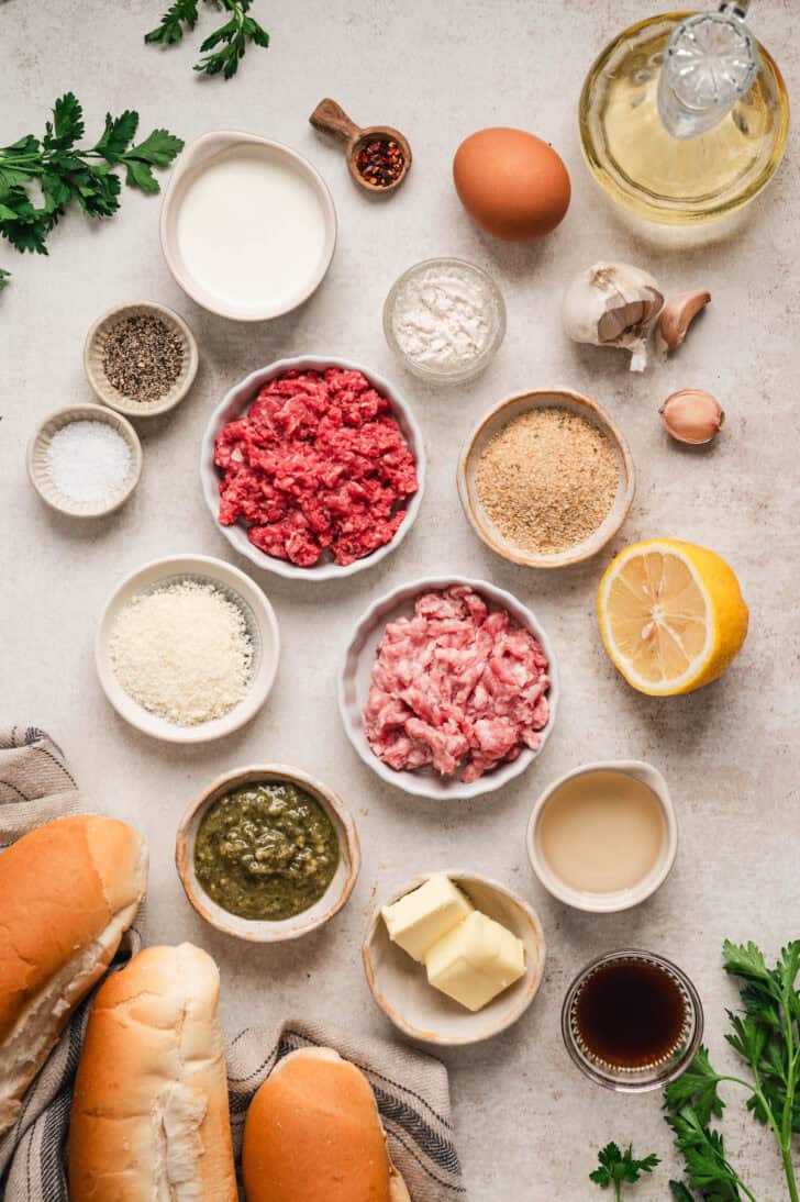 Ingredients on a light surface, including ground meat, breadcrumbs, cheese, spices, egg, pesto, butter, lemon, bread rolls and herbs.