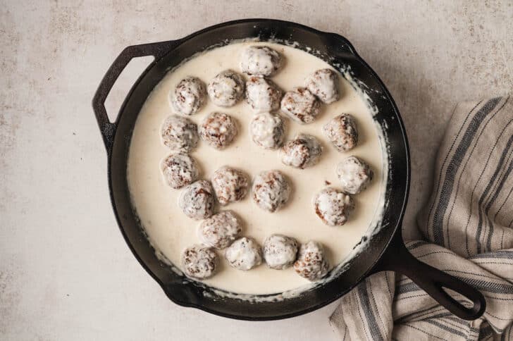 Meatballs for a meatball sandwich being cooked in cream sauce in a cast iron skillet.