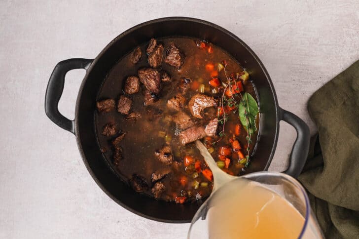 Broth being added to a meat and vegetable mixture in a Dutch oven.