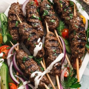 A large platter filled with kafta skewers, vegetables and a creamy dipping sauce.