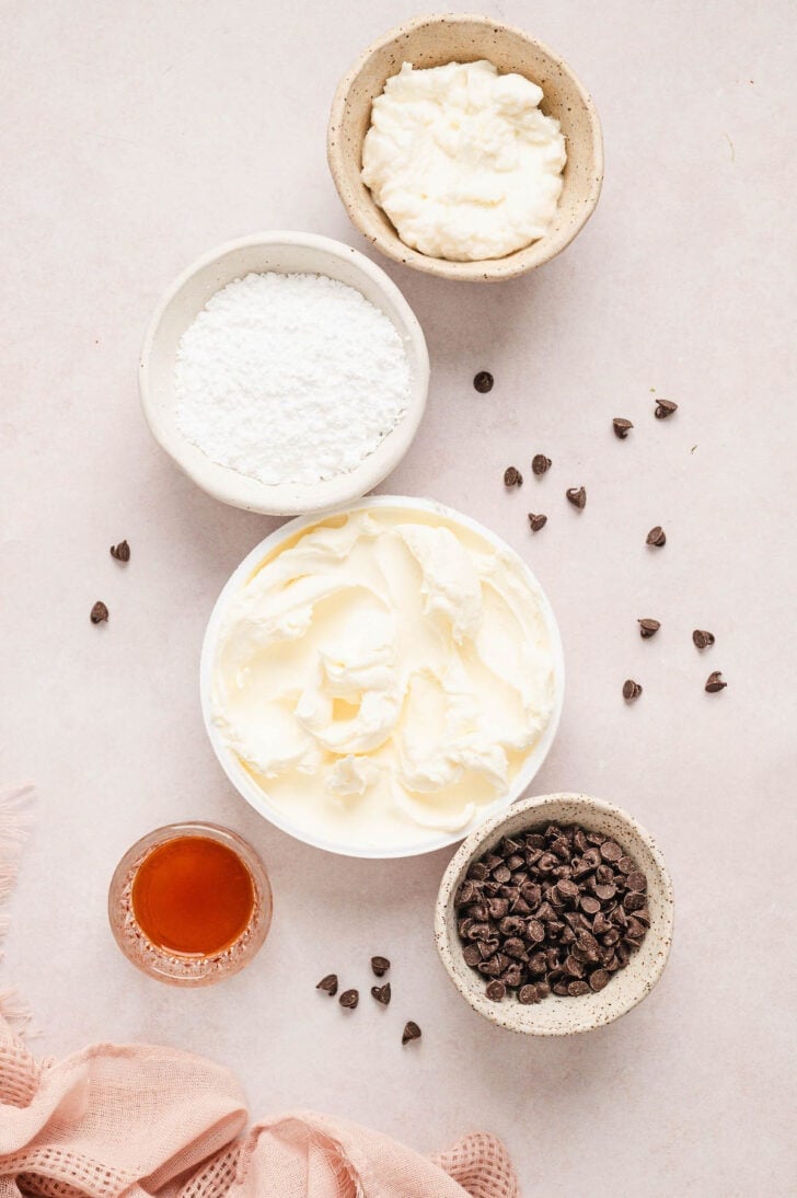 Ingredients on a light pink surface, including two types of soft cheese, powdered sugar, vanilla extract and mini chocolate chips.