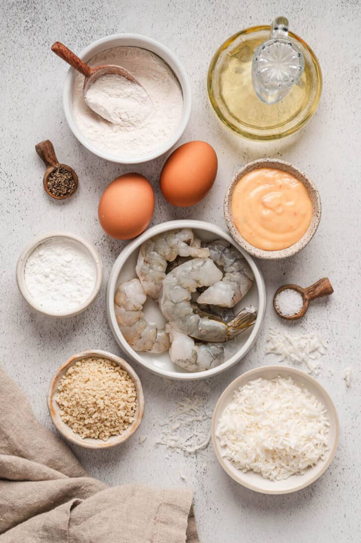 Ingredients on a white surface, including prawns, shredded dried fruit, breadcrumbs, flour, oil, eggs, spices and a dipping sauce.