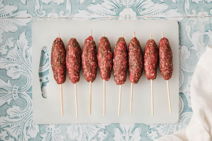 Raw meat kabobs on a white cutting board.