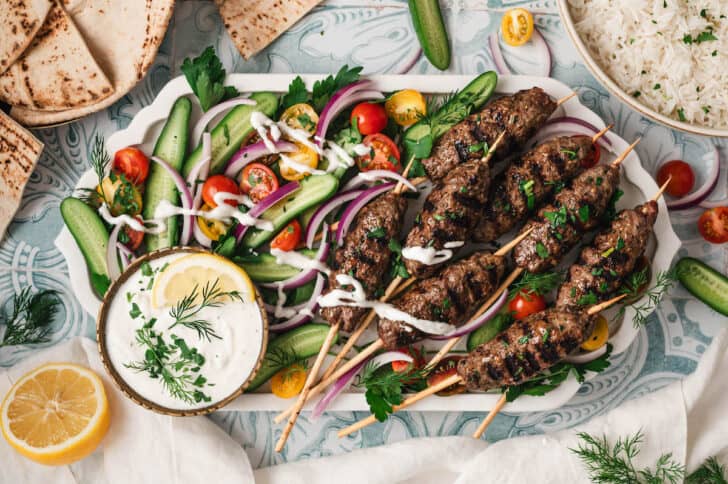 A large platter filled with beef kafta skewers, vegetables and a creamy dipping sauce.