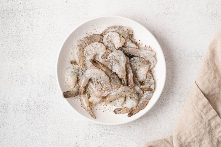 Raw shrimp sprinkled with salt and pepper in a white bowl.