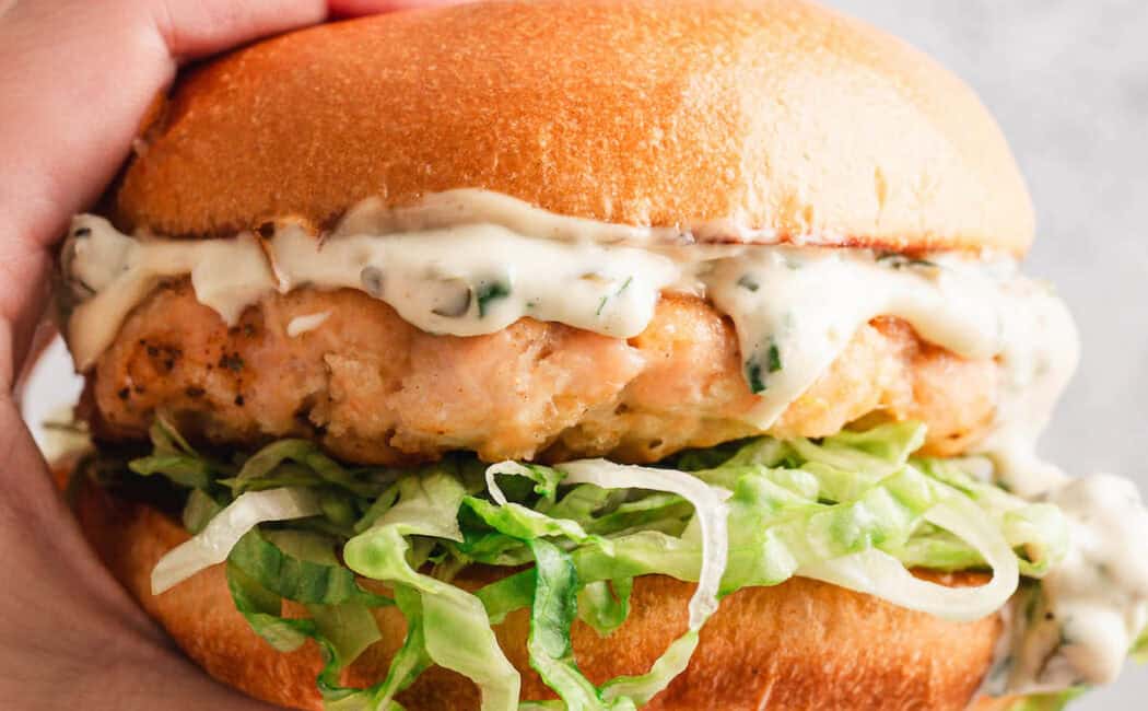 A hand holding a salmon burger in front of a light gray background.