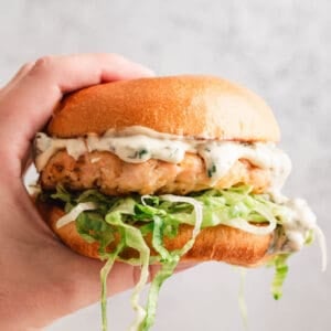 A hand holding a salmon burger in front of a light gray background.