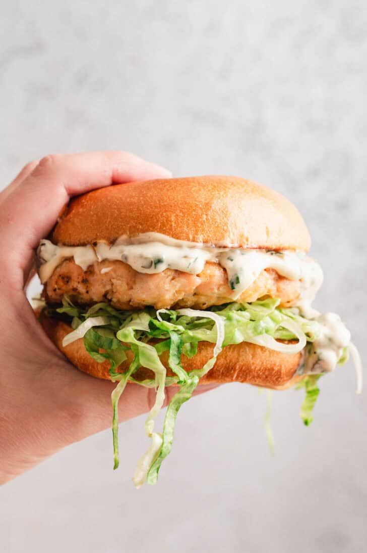 A hand holding a salmon burger in front of a light gray background.