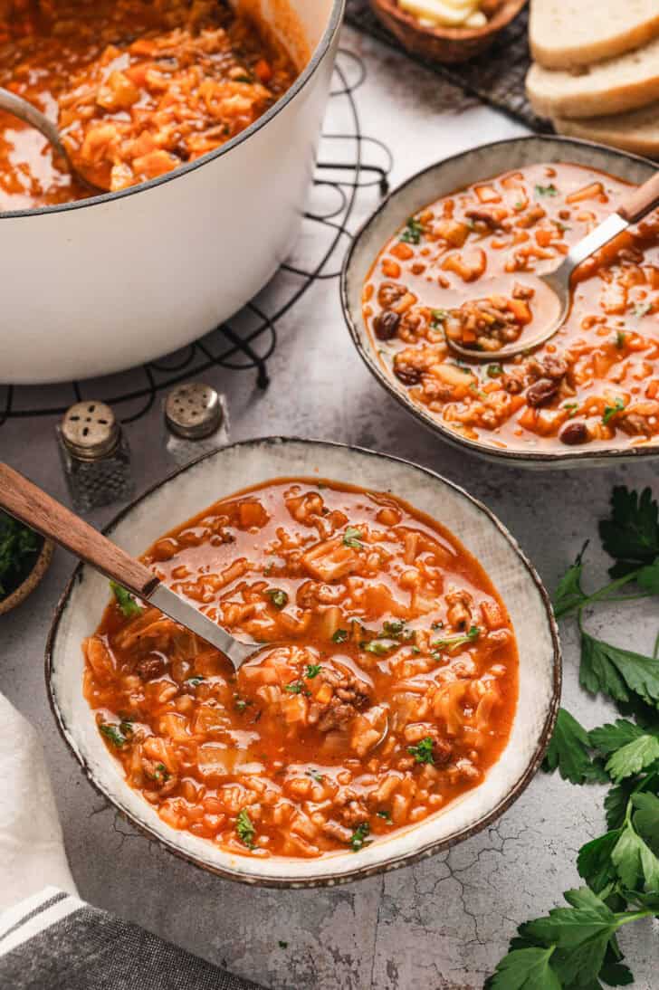 Two bowls of unstuffed cabbage roll soup with spoons in them, with the pot and sliced bread in the background.