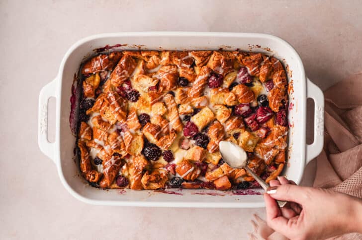 A hand spooning glaze over a breakfast casserole with croissants and berries.