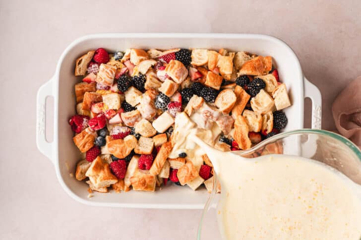 A white baking pan filled with cubed pastry pieces and berries, with a custard being poured over it.