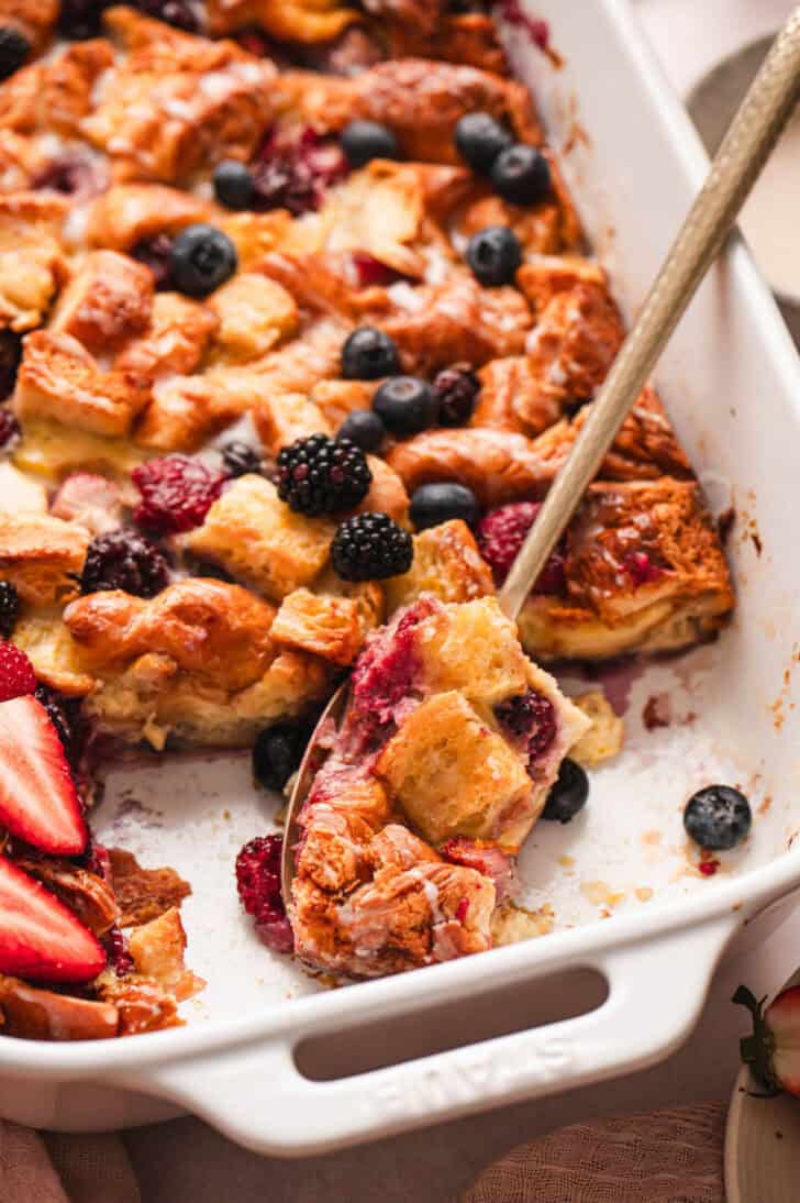 A spoon scooping a serving of croissant breakfast casserole from a white baking dish.