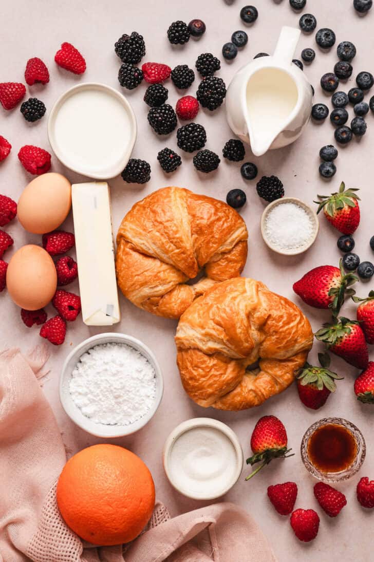 Ingredients on a light pink surface, including pastries, assorted berries, eggs, butter, sugar and dairy products.