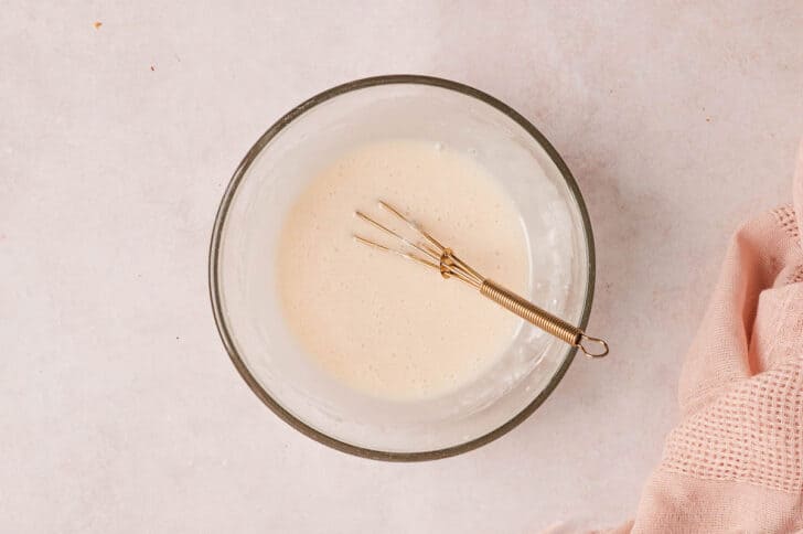 A glass bowl with a whisk mixing a white glaze in it.