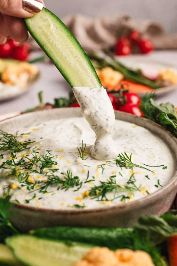 A cucumber spear being dipped into a bowl of homemade dill dip.