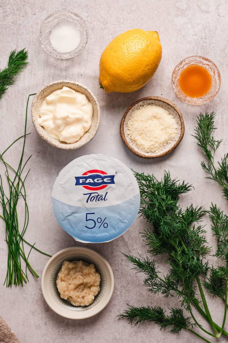 Ingredients on a textured surface, including Greek yogurt, mayonnaise, Parmesan cheese, horseradish, lemon and herbs.