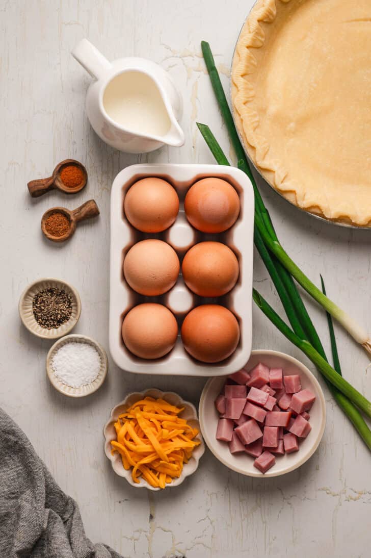 Ingredients on a light surface, including eggs, pie crust, ham, cheese, green onions, milk and spices.