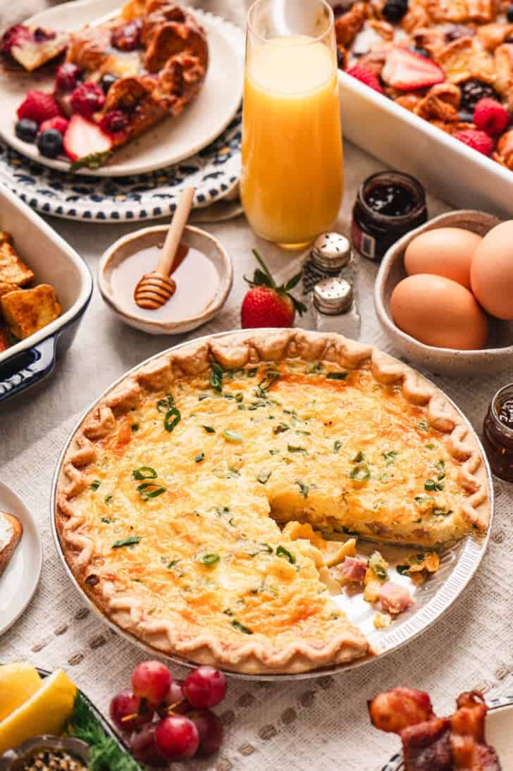 A ham quiche with one slice taken out of it in a brunch scene on a table.