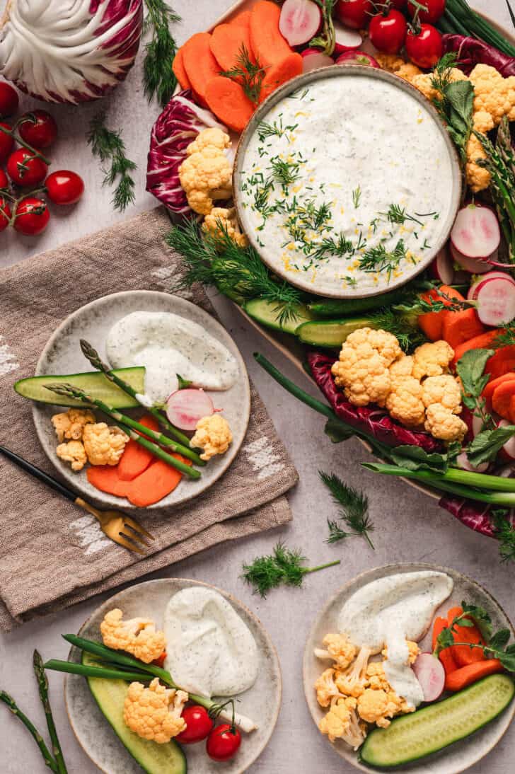 Three small plates filled with dill vegetable dip and various raw vegetables, with a big platter nearby.