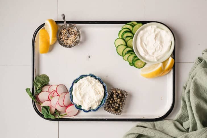 A white platter topped with bowls of creamy sauces, capers, spices and sliced veggies.