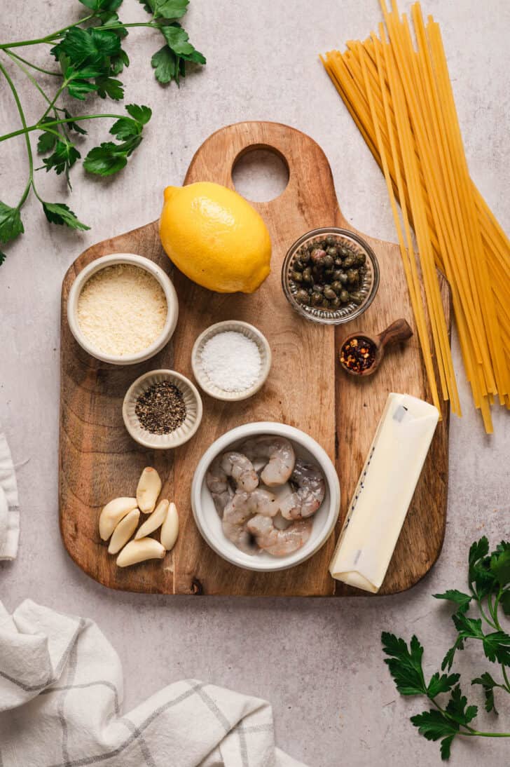 Ingredients on and around a wooden cutting board on a light background, including linguine, prawns, garlic, lemon, capers, butter, cheese and spices.