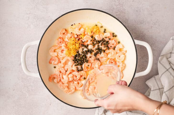 A hand pouring lemon juice into a white skillet with prawns and capers.