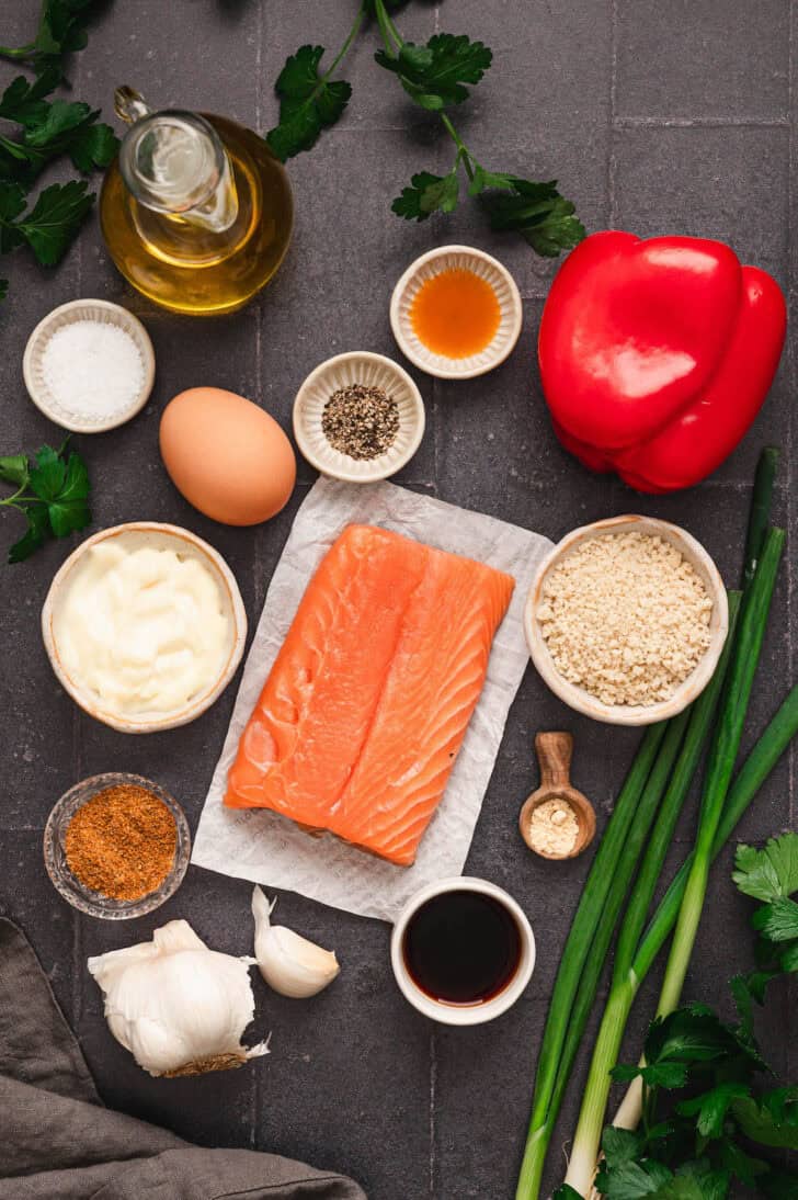 Ingredients on a dark surface, including raw sockeye fish, breadcrumbs, vegetables, spices, an egg, herbs and sauces.