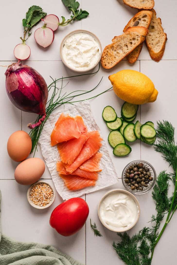 Ingredients on a white surface, including toasted bread, sliced veggies, creamy sauces, lox, eggs and capers.