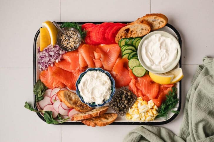 A white platter topped with bowls of creamy sauces, capers, spices, sliced veggies, lox, hard boiled eggs and toasted bread.