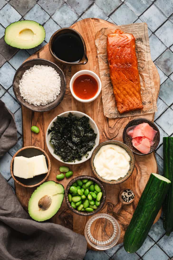 Ingredients on a wooden board on a tiled background, included smoked salmon, rice, avocado, herbs, and other sushi garnishes.