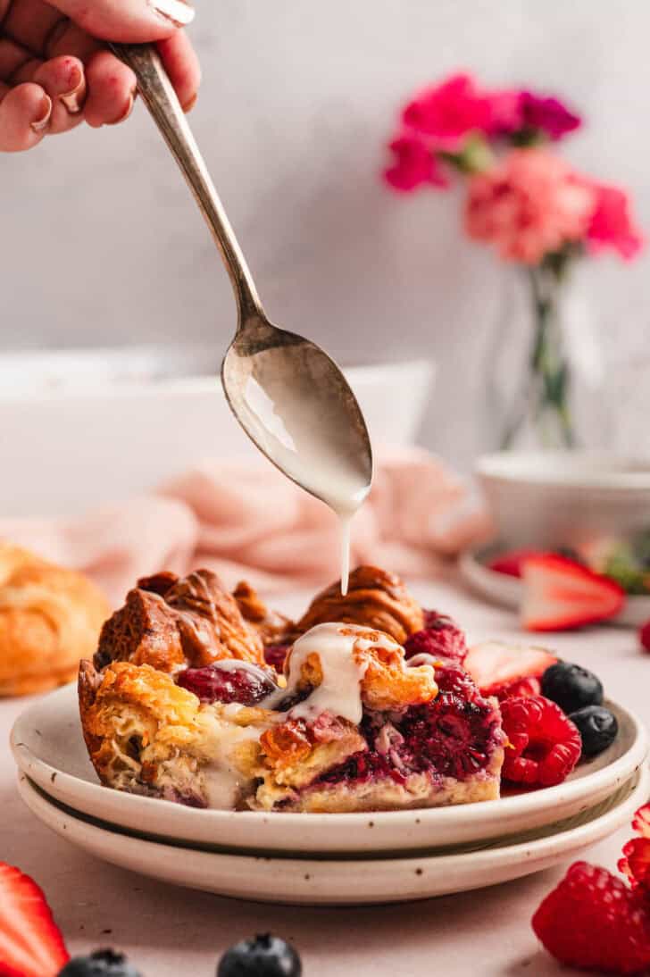 A hand spooning glaze over a piece of breakfast croissant casserole.