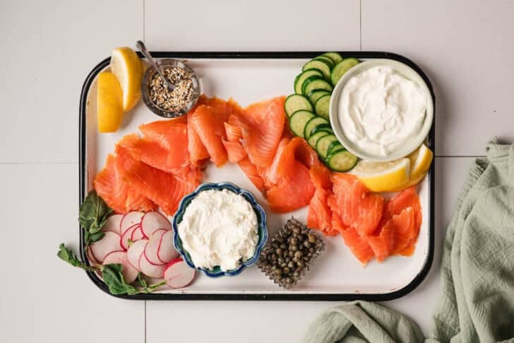 A white platter topped with bowls of creamy sauces, capers, spices, sliced veggies and lox.