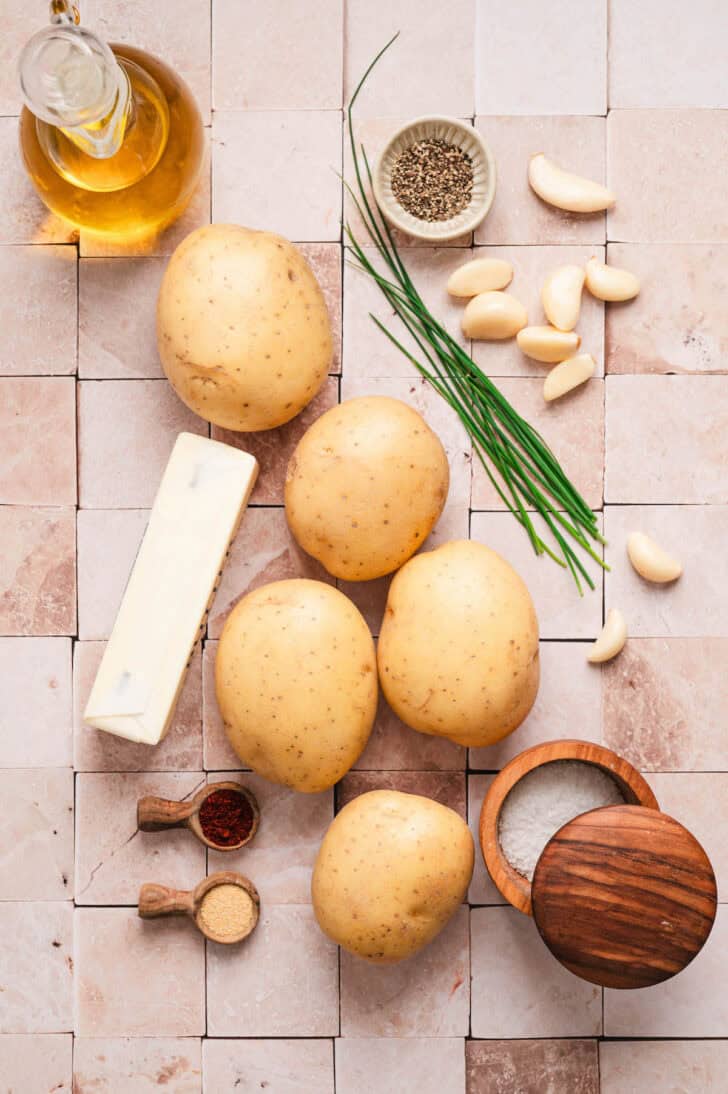 Ingredients on a tiled background, including yellow spuds, garlic, butter, spices and chives.