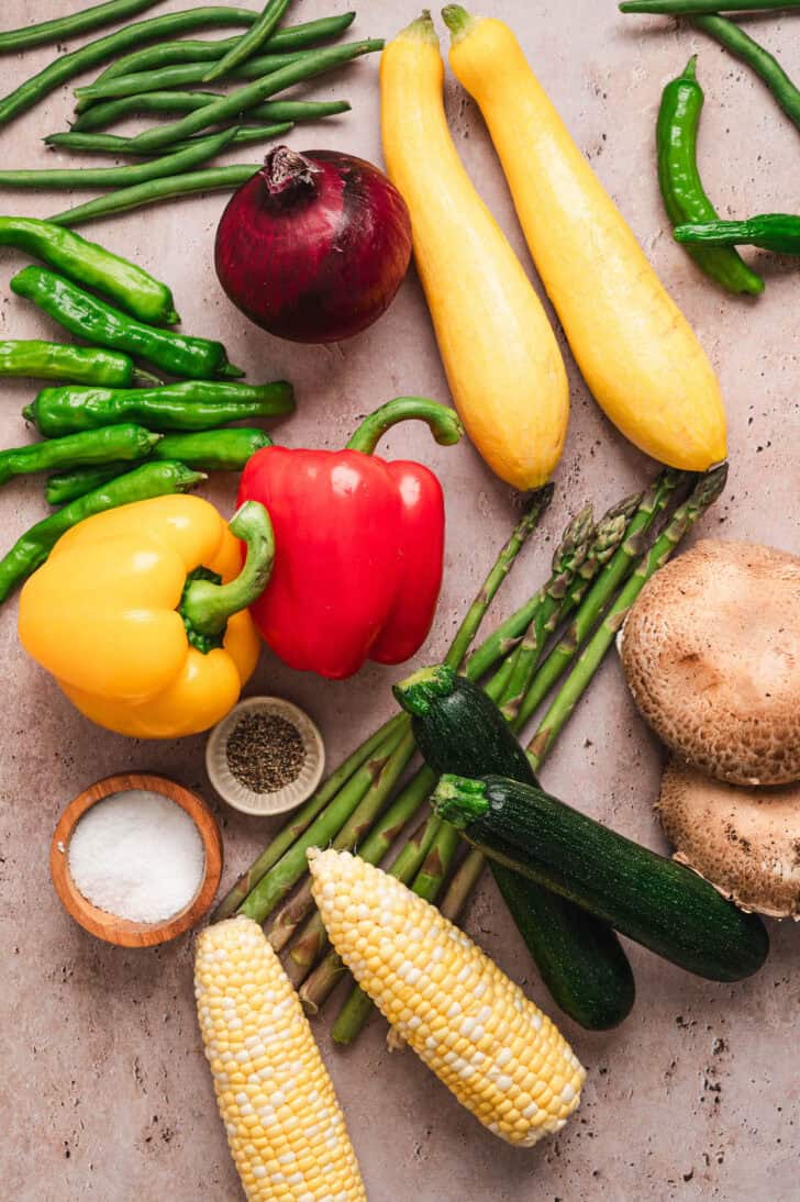 Veggies on a rustic background, including beans, asparagus, corn, bell peppers, squash, mushrooms, corn and onion.
