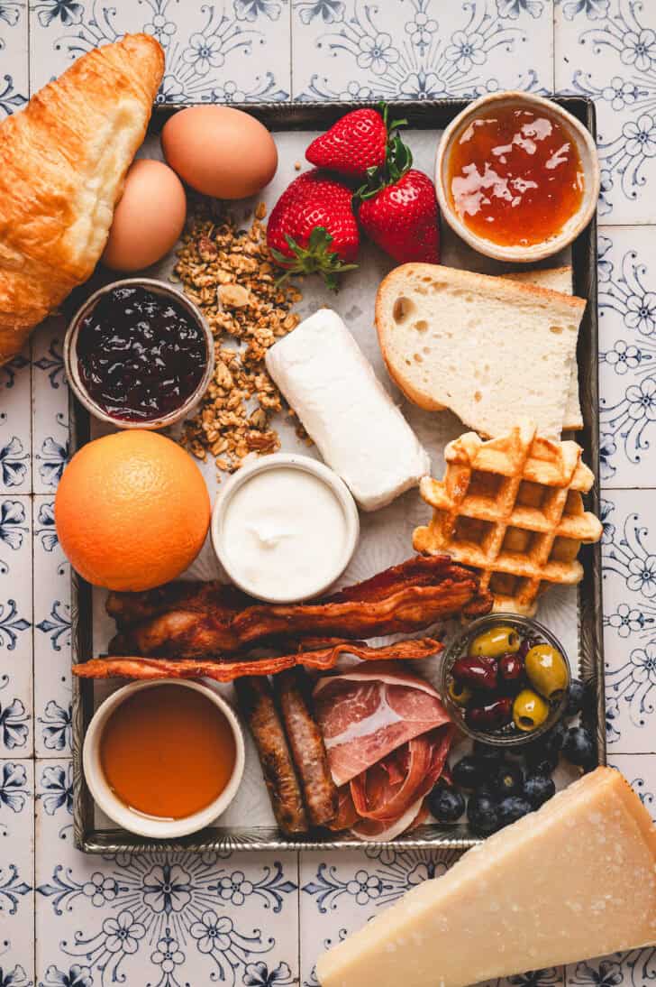 Ingredients on a rimmed baking pan, including bacon, sausage, prosciutto, cheeses, honey, fruit, granola, bread, waffles, eggs and olives.