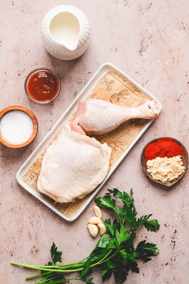 Ingredients on a rustic surface, including raw poultry, buttermilk, garlic, herbs and spices.